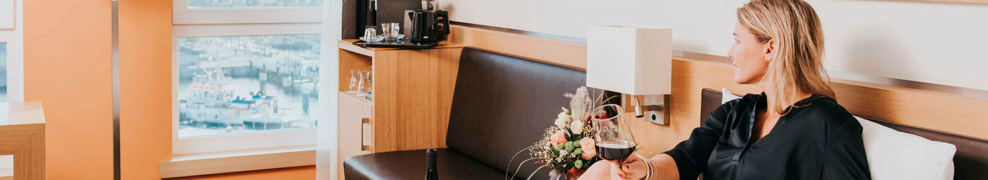 Innenansicht des Schlafzimmers der Suite mit Doppelbett und Förde-Aussicht im ATLANTIC Hotel Kiel Frau entspannt mit Weinglas auf Hotelbett, Blick aus Fenster auf Hafen, moderne Einrichtung, Blumenstrauß auf Tisch.