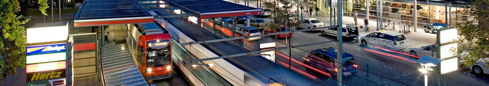 Außenansicht des Flughafens Bremen bei Abenddämmerung, mit Straßenbahn und vorbeifahrenden Autos.