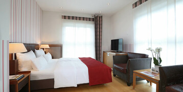 Helles Hotelzimmer mit Doppelbett, roten Akzenten, Sitzbereich, Flachbild-TV und großen Fenstern mit Vorhängen.