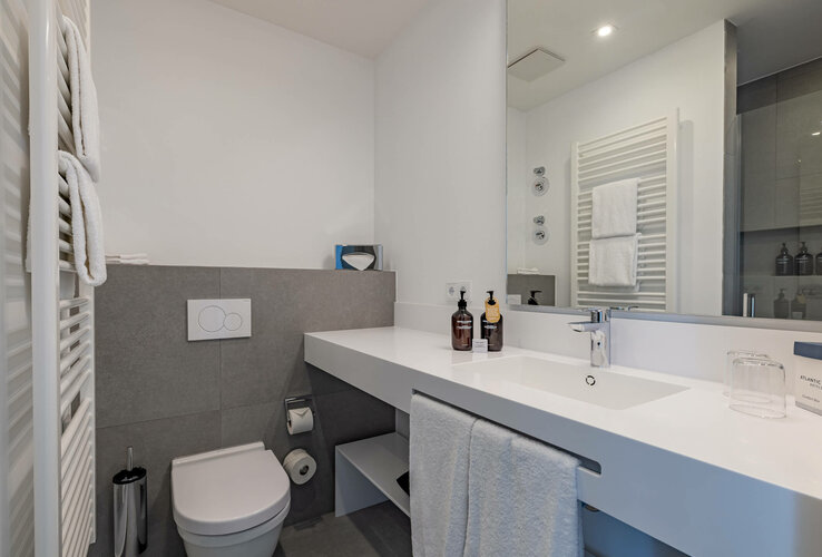 Modernes Badezimmer mit Waschbecken, Spiegel, Handtuchhalter, WC und Pflegeprodukten im ATLANTIC Grand Hotel Travemünde.