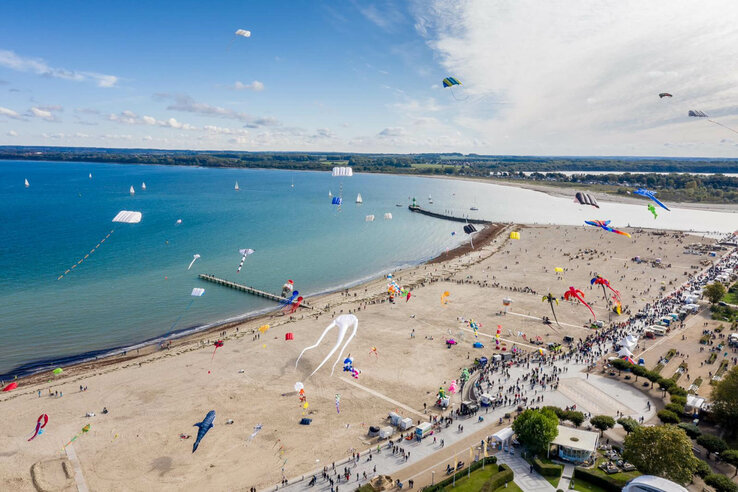 Luftaufnahme eines Strandes in Travemünde mit bunten Drachen am Himmel und vielen Menschen auf der Promenade.