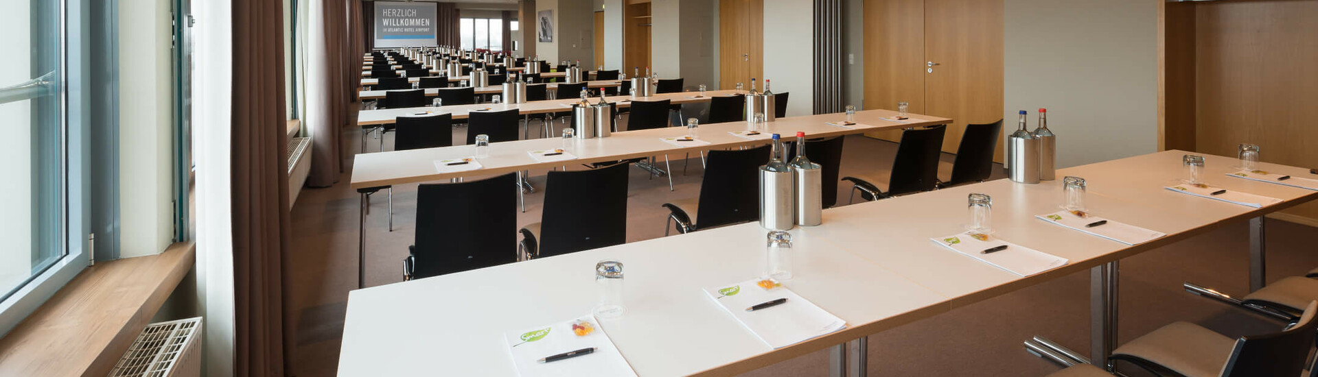 Veranstaltungsräume Lilienthal, Junkers, Lindbergh und Pazific im ATLANTIC Hotel Airport Bremen Heller Konferenzraum im ATLANTIC Hotel Airport Bremen, mit langen Tischen, Stühlen, Schreibmaterial und Flaschen.