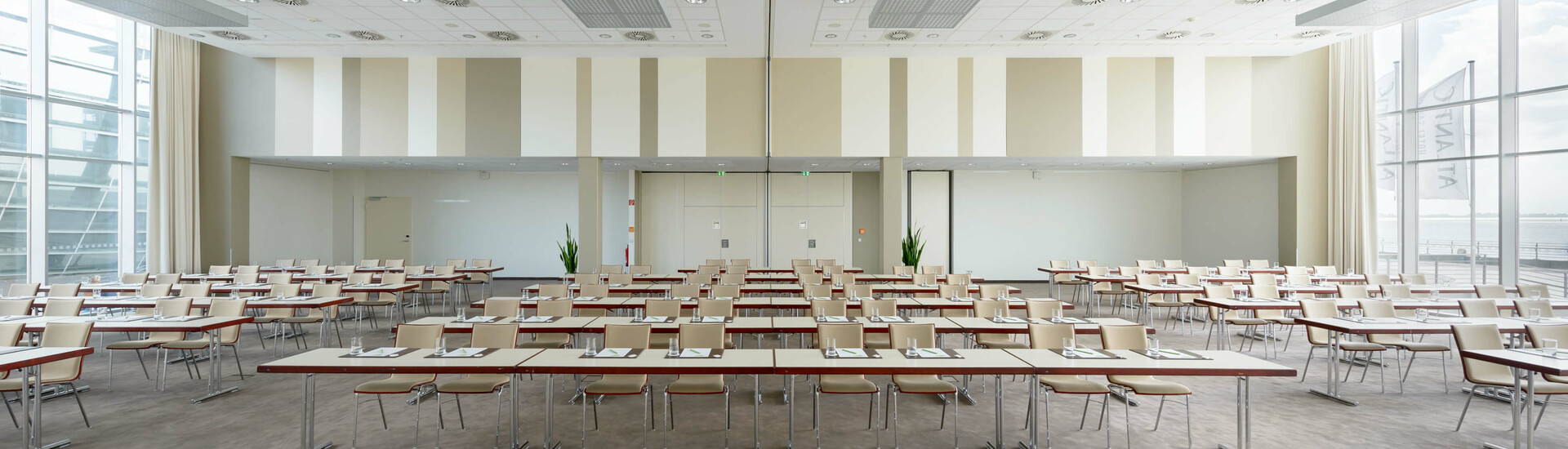 Parliamentary seating in conference room 1 and 2 at the ATLANTIC Hotel Sail City Bright conference room in the ATLANTIC Hotel Sail City, Bremerhaven, with rows of tables and chairs, large windows.