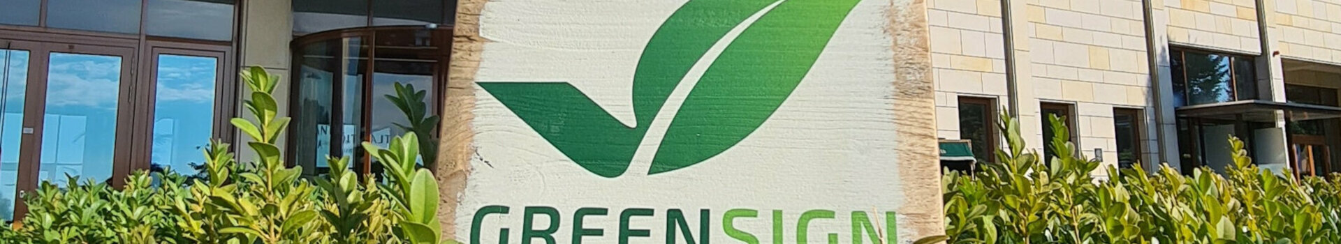 GreenSign Zertifikat | ATLANTIC Hotel Wilhelmshaven Fassade eines modernen Hotels mit "Greensign Hotel"-Schild und grünen Pflanzen im Vordergrund.