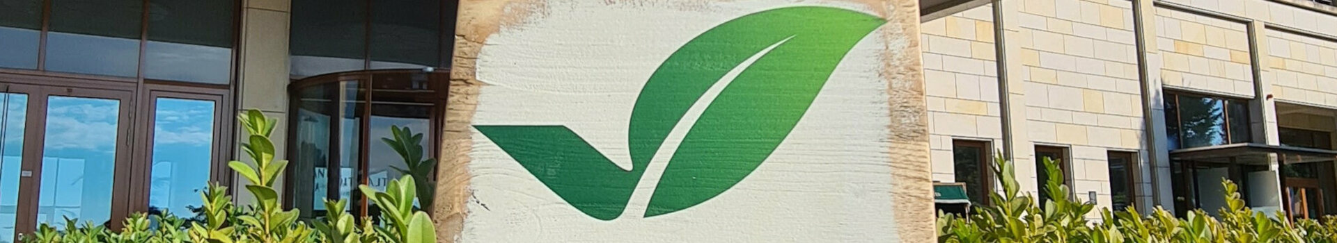 GreenSign Certificate | ATLANTIC Hotel Wilhelmshaven Facade of a modern hotel with "Greensign Hotel" sign and green plants in the foreground.