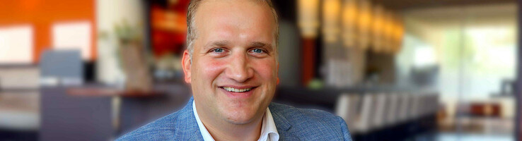 Interview mit Personaldirektor David Monte | ATLANTIC Hotels Ein lächelnder Mann in einem blauen Sakko sitzt in einer stilvollen Hotel-Lobby.