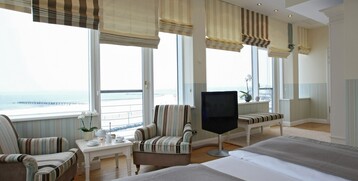 Bright hotel room with sea view, comfortable armchairs, table with tea service and large windows.