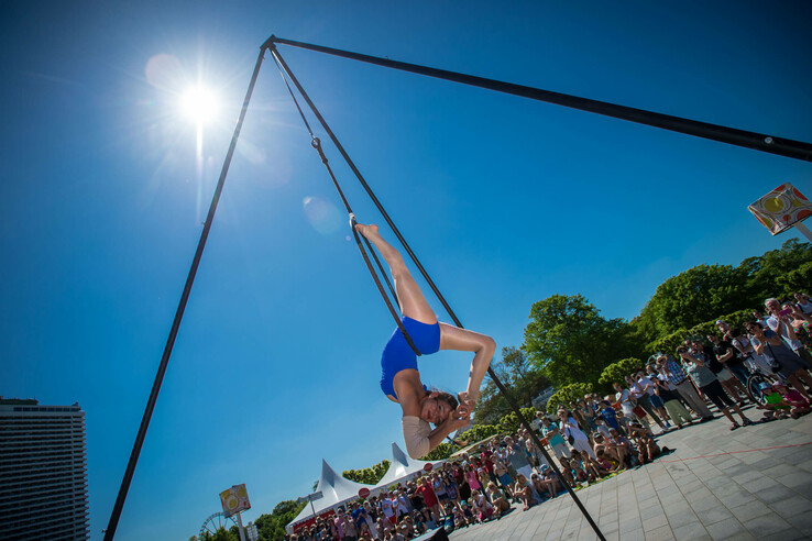 Akrobatin in blauer Kleidung schwebt kopfüber an einem Trapez unter freiem Himmel vor Publikum.