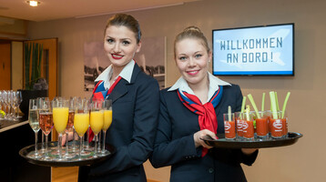 Zwei lächelnde Hostessen in Uniform servieren Sekt und Säfte; im Hintergrund ein "Willkommen an Bord"-Schild.
