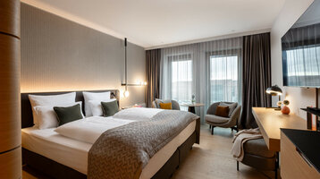 Modern hotel room with double bed, desk, two armchairs and large windows with curtains at the ATLANTIC Hotel Heidelberg.