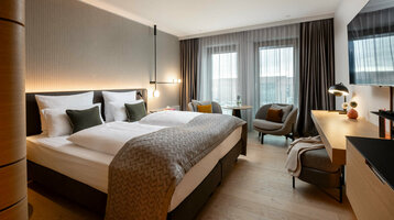 Modernes Hotelzimmer mit Doppelbett, Schreibtisch, zwei Sesseln und großen Fenstern mit Vorhängen im ATLANTIC Hotel Heidelberg.