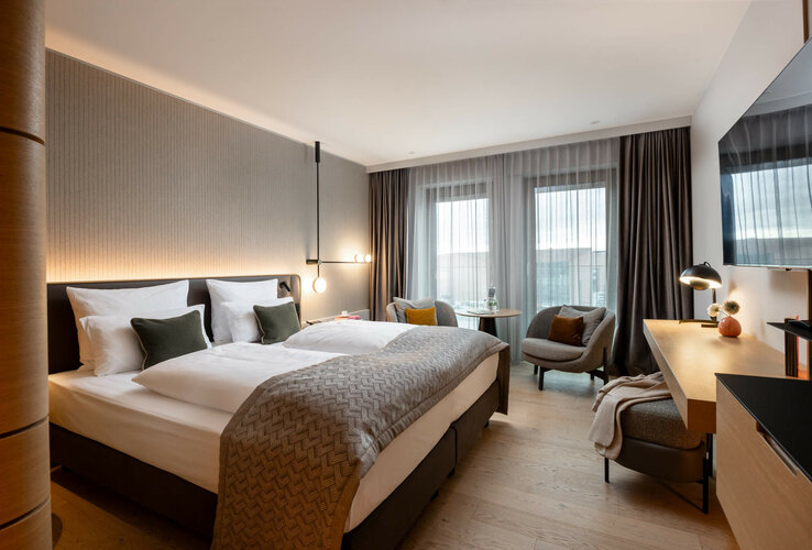 Modernes Hotelzimmer mit Doppelbett, Schreibtisch, zwei Sesseln und großen Fenstern mit Vorhängen im ATLANTIC Hotel Heidelberg.