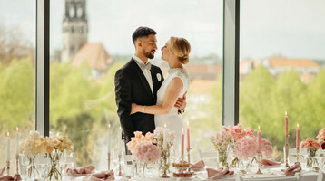 Brautpaar vor großer Fensterfront, elegant gedeckter Tisch mit Blumen und Kerzen, Ausblick auf Münster.