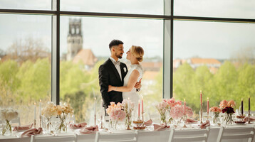 Brautpaar vor großer Fensterfront, elegant gedeckter Tisch mit Blumen und Kerzen, Ausblick auf Münster.