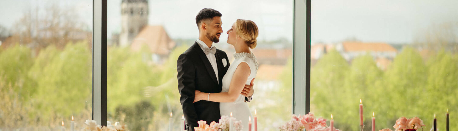Himmlisch Heiraten im Engelsaal des ATLANTIC Hotel Münster Brautpaar vor großer Fensterfront, elegant gedeckter Tisch mit Blumen und Kerzen, Ausblick auf Münster.