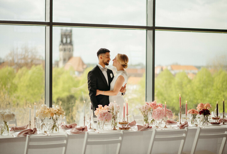 Brautpaar vor großer Fensterfront, elegant gedeckter Tisch mit Blumen und Kerzen, Ausblick auf Münster.