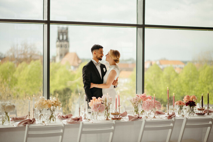 Himmlisch Heiraten im Engelsaal des ATLANTIC Hotel Münster Brautpaar vor großer Fensterfront, elegant gedeckter Tisch mit Blumen und Kerzen, Ausblick auf Münster.