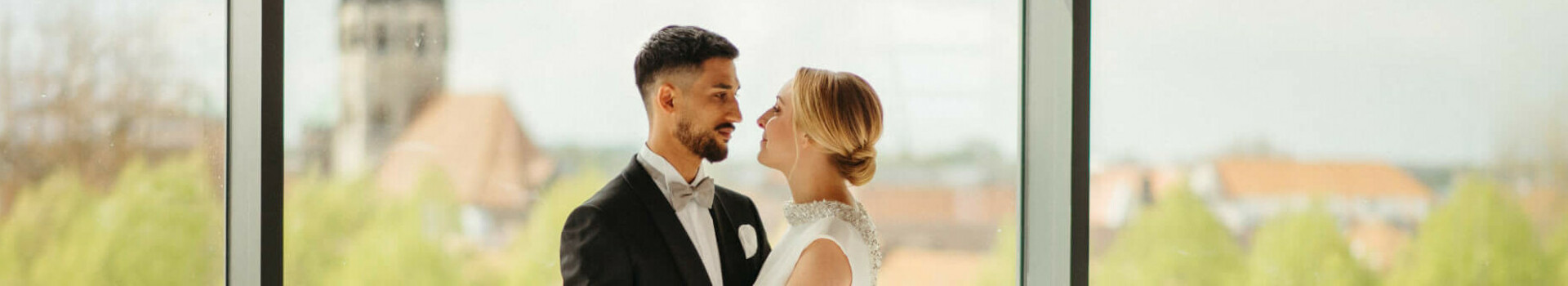 Brautpaar vor großer Fensterfront, elegant gedeckter Tisch mit Blumen und Kerzen, Ausblick auf Münster.