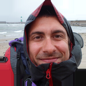 Eine Person in Outdoor-Kleidung lächelt am Strand, im Hintergrund ist ein Leuchtturm zu sehen.