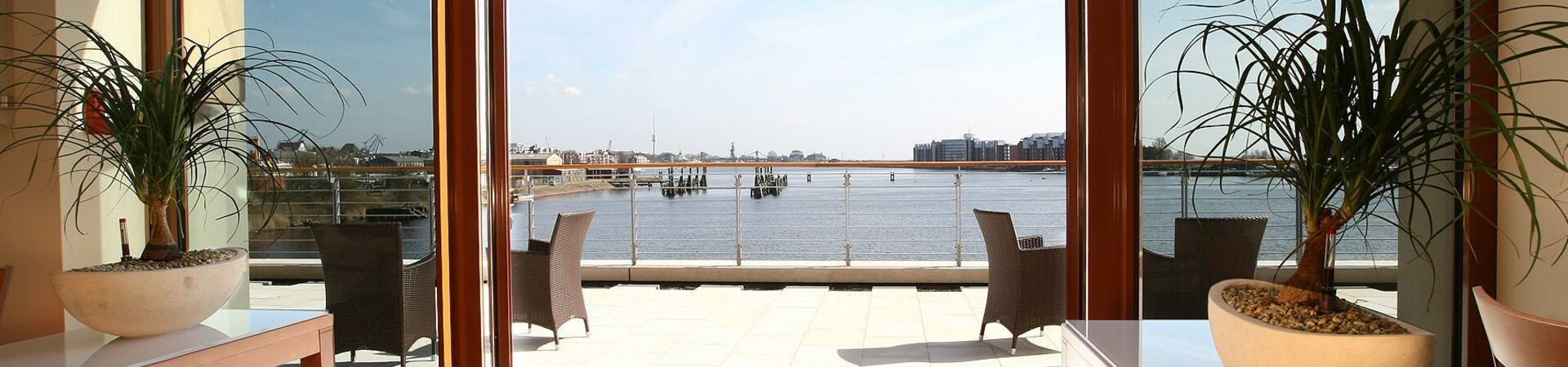 Ausblick auf den Hafen vom Restaurant Harbour View des ATLANTIC Hotel Wilhelmshaven Helle Terrasse mit Blick aufs Wasser, modernen Möbeln und Pflanzen im Vordergrund.