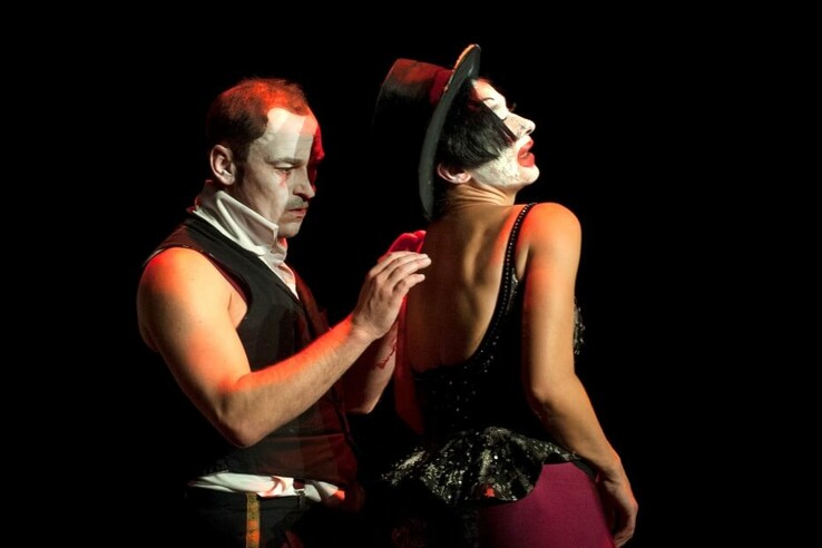 Two artists in circus costumes with made-up faces on a dark stage, one wearing a hat. © bremer shakespeare company