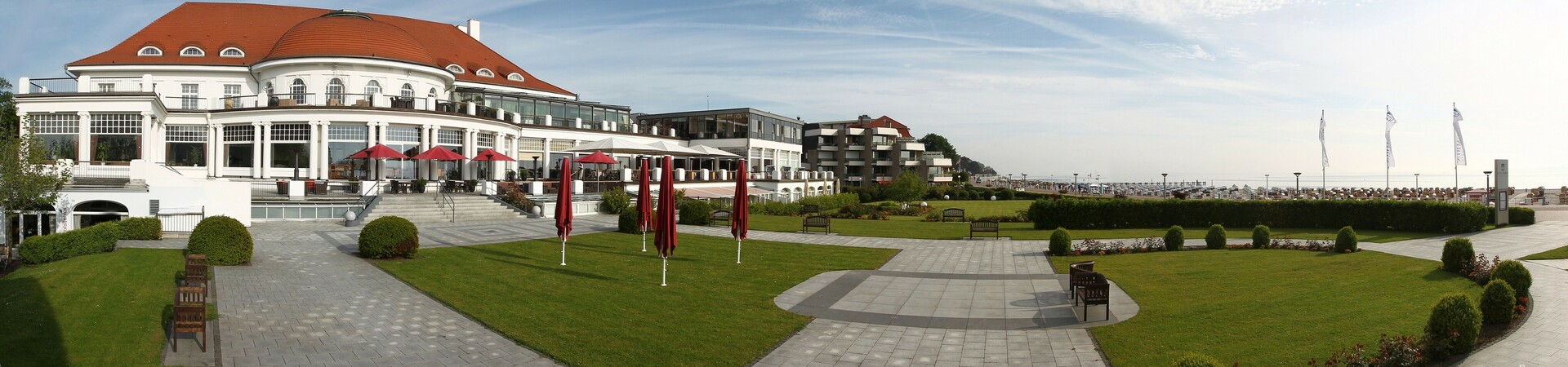ATLANTIC Grand Hotel Travemünde mit gepflegtem Garten, roten Sonnenschirmen und Blick auf die Ostsee.