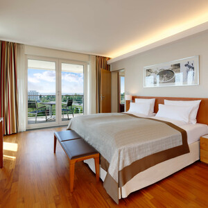 Executive Juniorsuite im ATLANTIC Hotel Wilhelmshaven Helles Hotelzimmer mit Doppelbett, Holzboden, TV, Balkon mit Stühlen und weitem Ausblick auf grüne Landschaft.