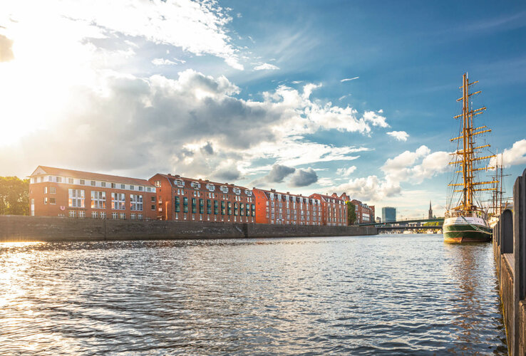 View of the Weser with a sailing ship and red buildings in the background, under a blue sky with clouds.