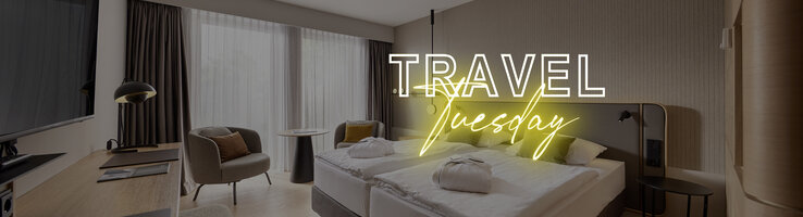 Modernes Hotelzimmer mit Doppelbett, Schreibtisch, Sessel und großen Fenstern, Text "Travel Tuesday" in Neon.