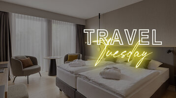 Modernes Hotelzimmer mit Doppelbett, Schreibtisch, Sessel und großen Fenstern, Text "Travel Tuesday" in Neon.