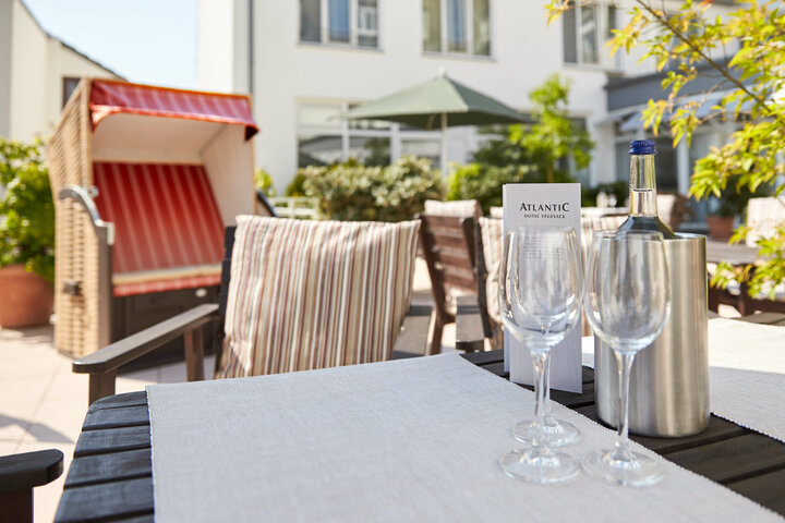Genießen Sie die Sonne in unserem Innenhof mit Terrasse in Vegesack, Bremen Terrasse des ATLANTIC Hotel Vegesack mit Strandkorb, Tisch, Gläsern und Flasche, umgeben von grünen Pflanzen.