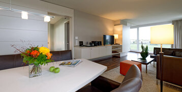 Modern hotel suite with leather armchairs, dining table, bouquet of flowers, flat-screen TV and view of the racecourse through large windows.