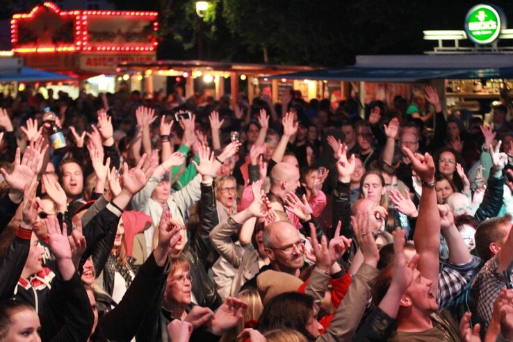 Crowd at a festival evening, many raising their hands, illuminated stands in the background.