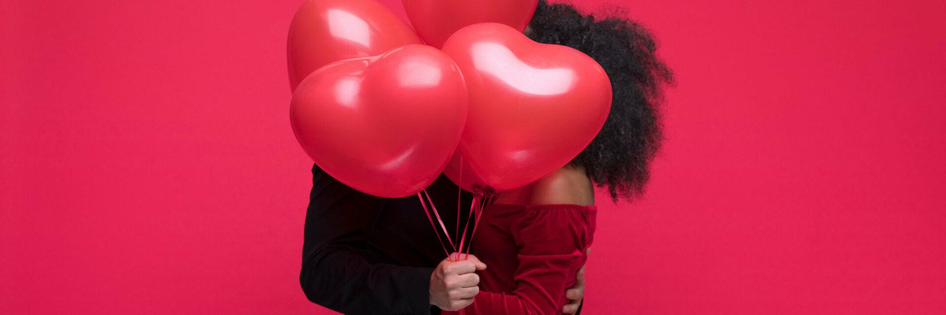 Couple hugging behind red heart balloons against a red background.