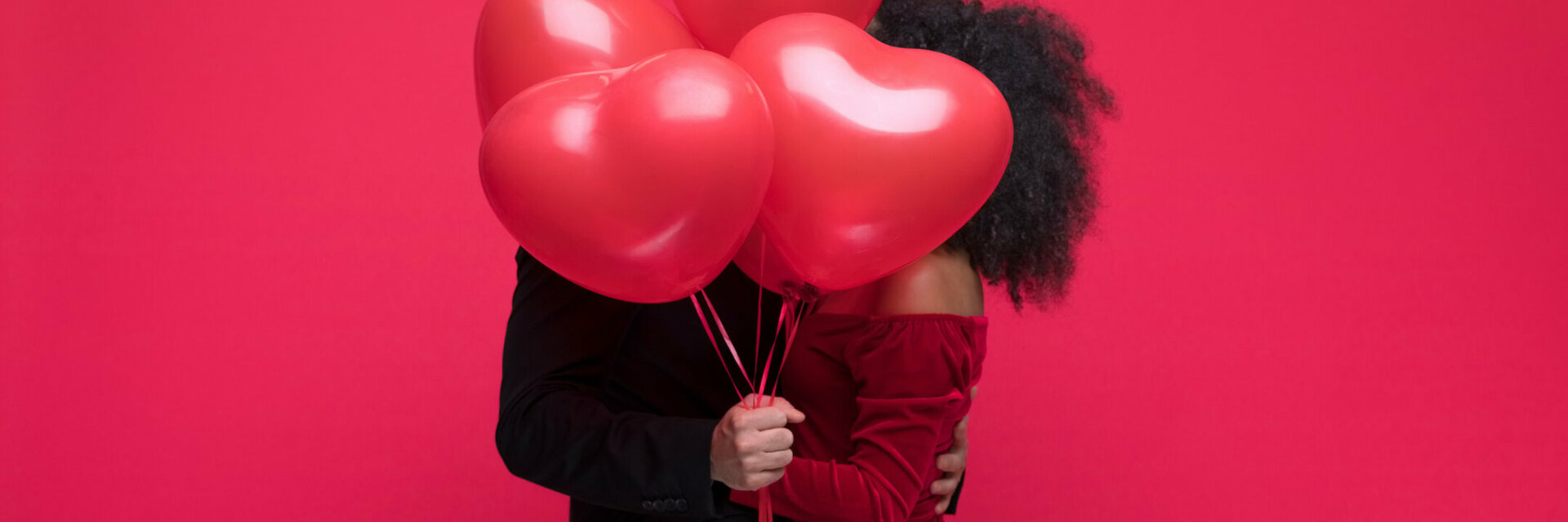 Paar umarmt sich hinter roten Herzluftballons vor rotem Hintergrund.