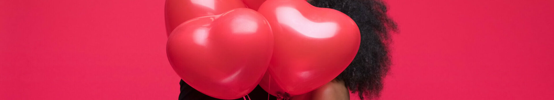 Paar umarmt sich hinter roten Herzluftballons vor rotem Hintergrund.