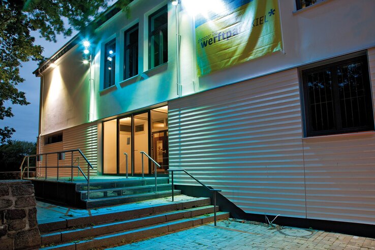 Eingangsbereich eines modern beleuchteten Gebäudes mit Glasfront und Treppen bei Nacht.