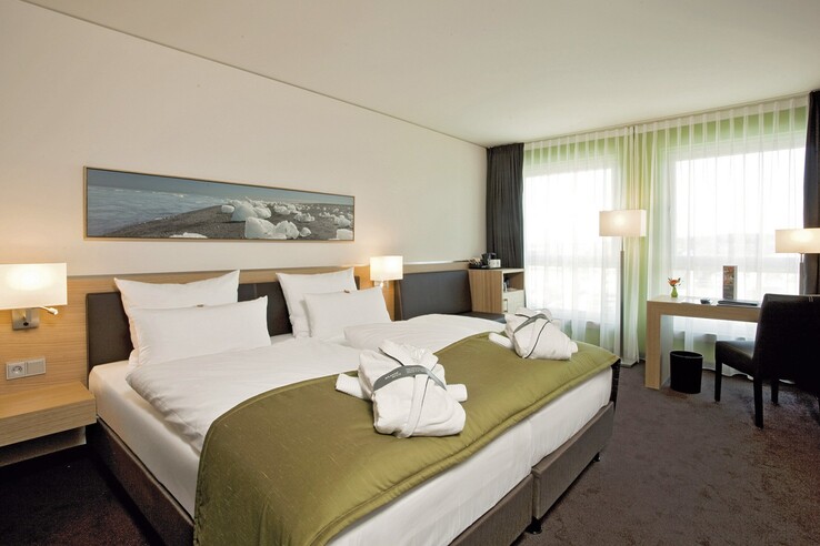 Modernes Hotelzimmer mit Doppelbett, grüner Tagesdecke, Schreibtisch, Sessel und großen Fenstern im ATLANTIC Hotel Kiel.