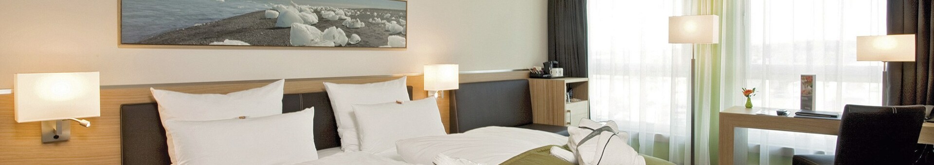 Modernes Hotelzimmer mit Doppelbett, grüner Tagesdecke, Schreibtisch, Sessel und großen Fenstern im ATLANTIC Hotel Kiel.