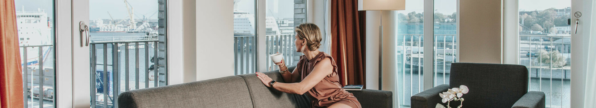 Living area with a couch, a table and flowers in the suite at the ATLANTIC Hotel Kiel Bright hotel suite with sofa and view of the harbor through large windows in the ATLANTIC Hotel Kiel.
