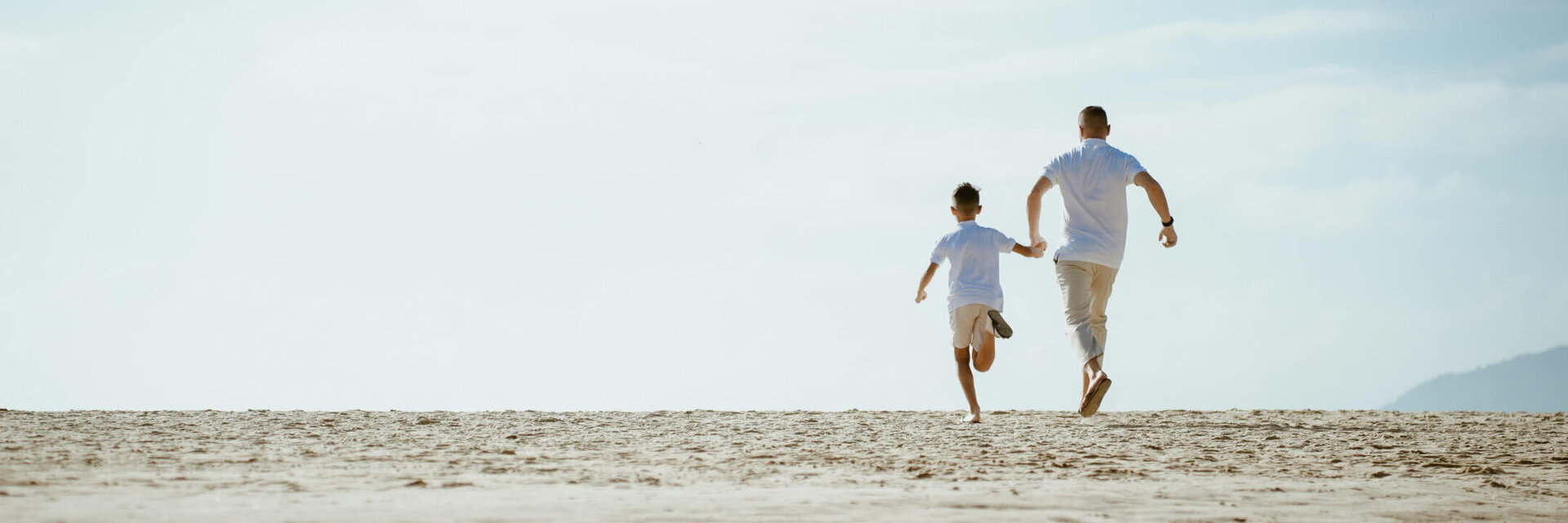 Zwei Personen laufen fröhlich am Sandstrand entlang, blauer Himmel im Hintergrund.