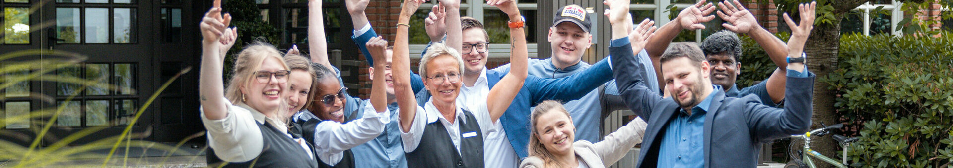 Gruppe von Hotelmitarbeitern vor dem Eingang, lächelnd und winkend, umgeben von Pflanzen.