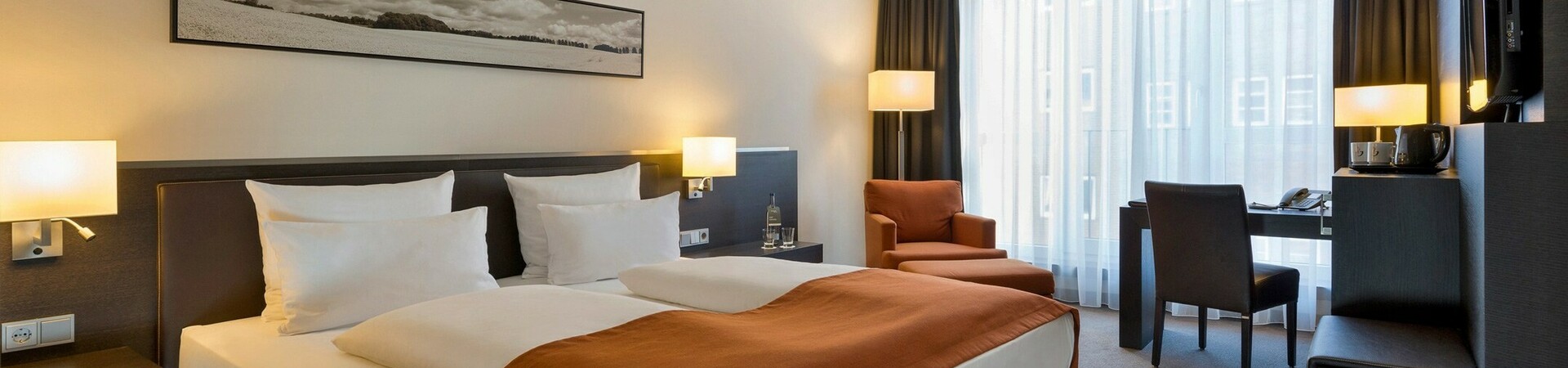Innenansicht des Comfort Zimmers im ATLANTIC Hotel Lübeck Modernes Hotelzimmer mit Doppelbett, Schreibtisch, Sessel und großen Fenstern mit Vorhängen im ATLANTIC Hotel Lübeck.