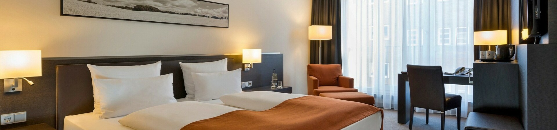 Modern hotel room with double bed, desk, armchair and large windows with curtains at the ATLANTIC Hotel Lübeck.