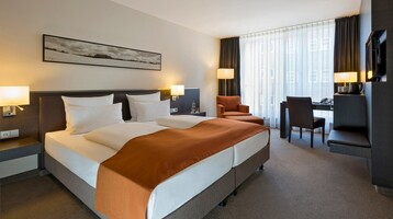 Innenansicht des Comfort Zimmers im ATLANTIC Hotel Lübeck Modernes Hotelzimmer mit Doppelbett, Schreibtisch, Sessel und großen Fenstern mit Vorhängen im ATLANTIC Hotel Lübeck.