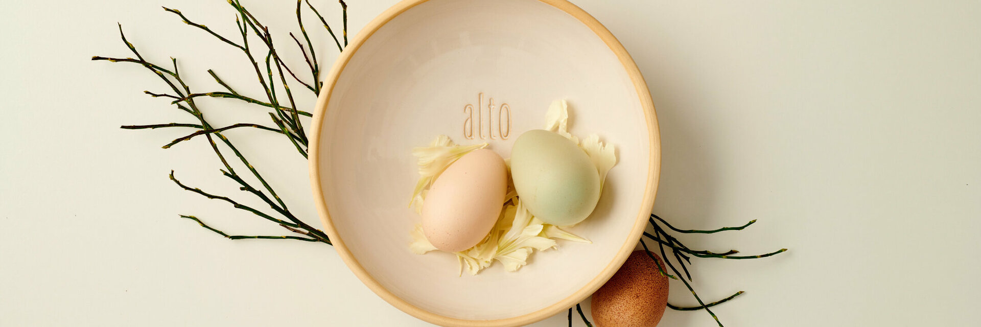 Zwei bunte Eier in einer Schale mit Blütenblättern, dekoriert mit Zweigen, Schriftzug "alto".