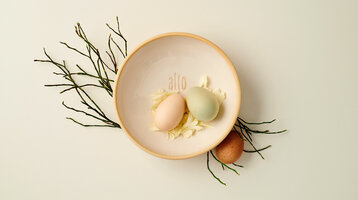 Zwei bunte Eier in einer Schale mit Blütenblättern, dekoriert mit Zweigen, Schriftzug "alto".