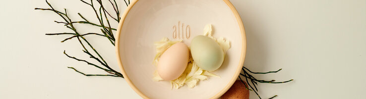 Zwei bunte Eier in einer Schale mit Blütenblättern, dekoriert mit Zweigen, Schriftzug "alto".