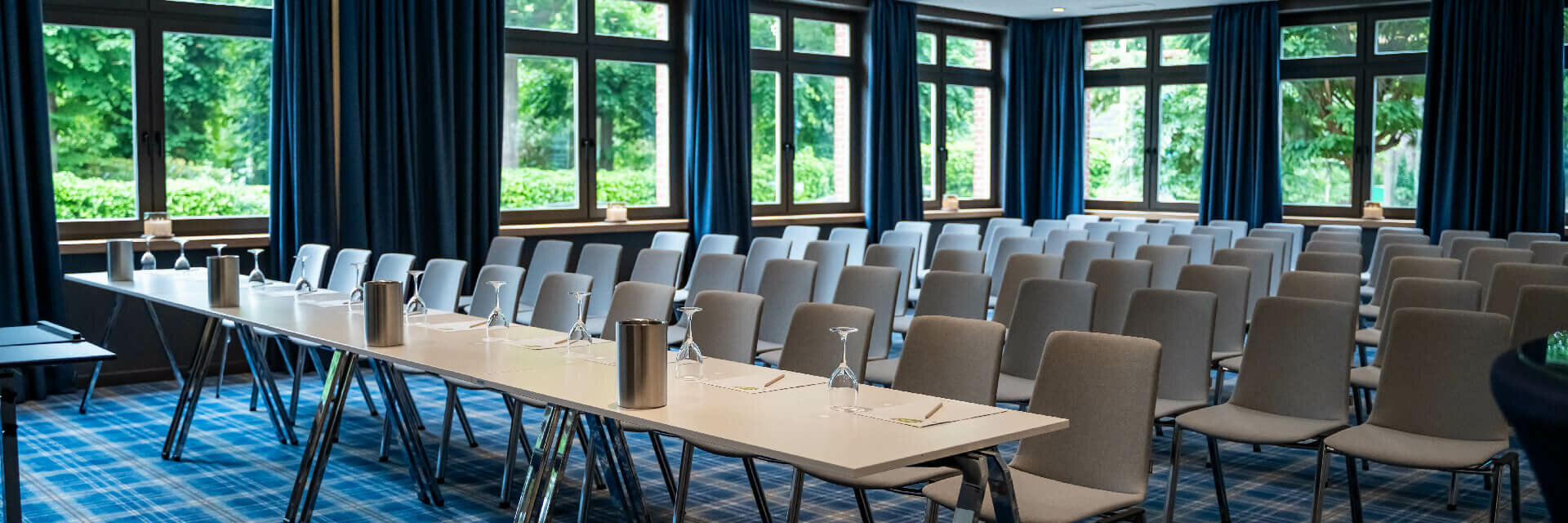 Moderner Konferenzraum im ATLANTIC Hotel mit blauen Vorhängen, Fensterfront, grauen Stühlen und gedecktem Tisch.