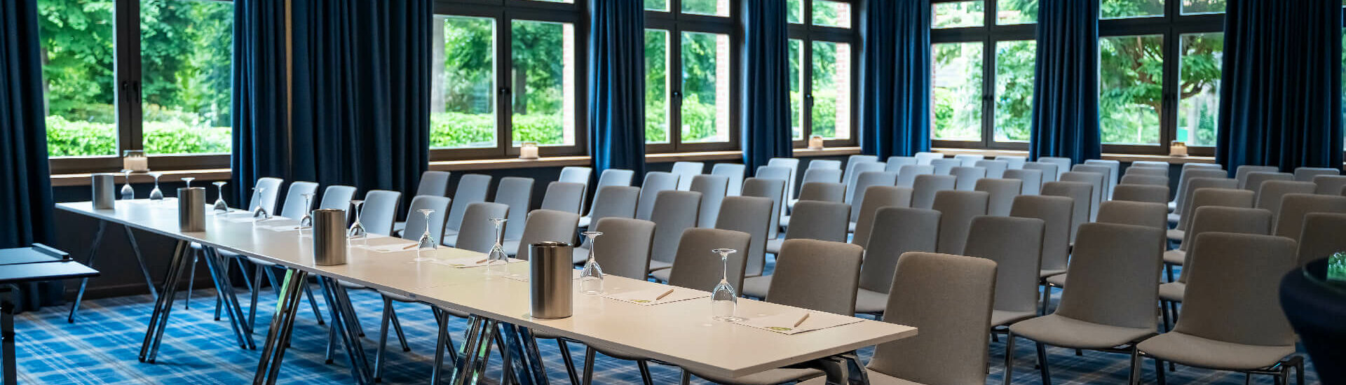 Moderner Konferenzraum im ATLANTIC Hotel mit blauen Vorhängen, Fensterfront, grauen Stühlen und gedecktem Tisch.